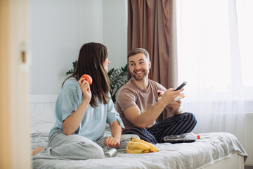 Fototapeta na wymiar Happy young man and woman relaxing on bed at home watching TV together and eating donuts and fruit. The concept of rest and junk food.
