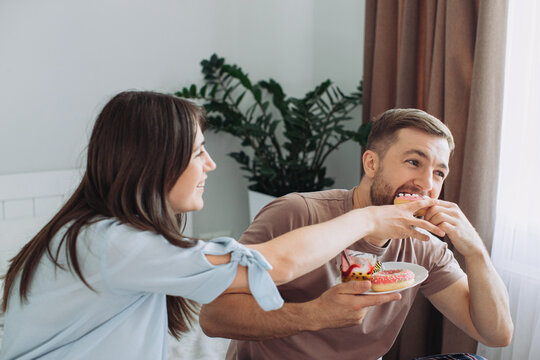 Happy young man and woman relaxing on bed at home watching TV together and eating donuts and fruit. The concept of rest and junk food.