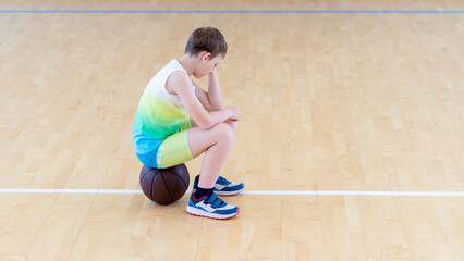 Sad disappointed boy sitting on basketball ball in a physical education lesson