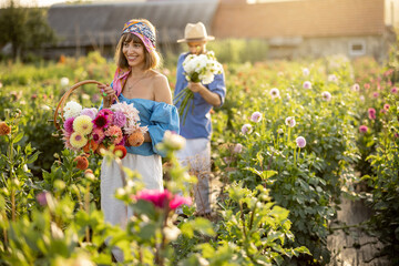 Man and a woman pick up dahlia flowers while working at rural flower farm with farmhouse on...