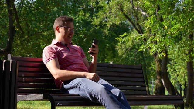 Attractive millennial male in glasses celebrating win on smartphone, sitting on bench at city park outdoors. Happy man checking news on smart phone