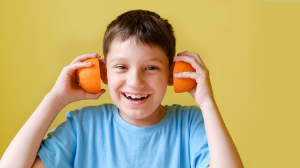 boy with orange. A smiling boy in a blue shirt stands on a yellow background and holds orange halves near his ears, instead of headphones, as if listening to music.