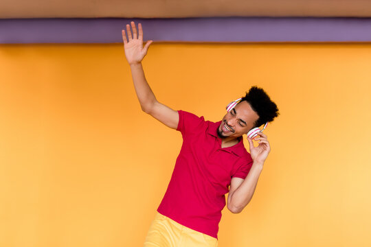 Exited young afro-american guy with happy emotions closed eyes, holding up hand and dancing while listening music and having fun over yellow background 