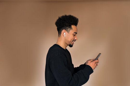 Profile portrait of attractive man dark-skinned young guy with wavy black hair wearing black shirt listening music ad using smartphone posing over beige background 