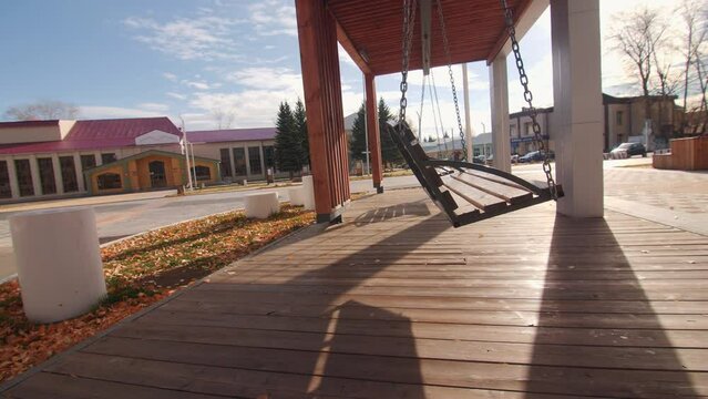 Empty Wooden Porch Swing In Arbor Installed In Modern Urban Park. Stylish Hanging Bench In Public City Garden At Sunset