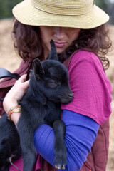 Close up of female with hat on hugging a very young goat in her arms.