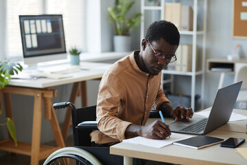 Portrait of black young man with disability working in office and using laptop, accessible...