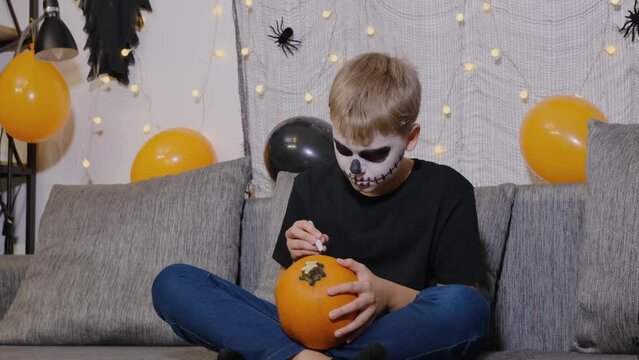 A funny child with makeup on his face is preparing for the celebration of Halloween by painting a pumpkin.