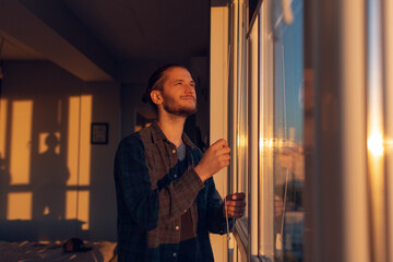 Portrait of young smiling man closes window rolled jalousies in sunny day at sunset.