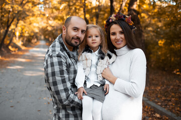 Portrait of happy family on background of golden park in autumn day. Father, daughter and pregnant mother.