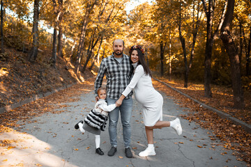 Portrait of happy family on background of golden park in autumn day. Father, daughter and pregnant mother.