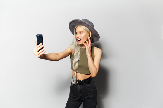 Studio portrait of cute happy blonde girl while making selfie photo and listen the music via wireless earbuds, holding smartphone in hand, wearing grey hat on white background.