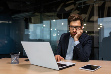 sad and depressed businessman in depression working inside modern office building, man in business suit with glasses and beard using laptop for work, boss disappointed with work result.