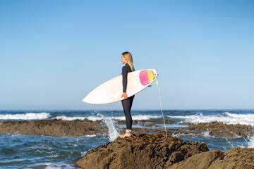 Surfer girl at the beach standing on a rock with her surfboard in the morning. Female surfer - Powered by Adobe