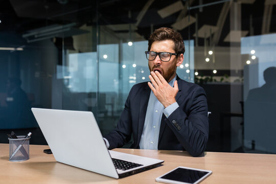 Mature bored businessman working inside modern office building, boss in glasses and business suit using laptop, investor yawning sitting on chair at table. - Powered by Adobe