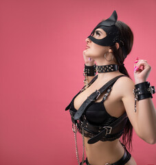 brunette in bdsm leather straps on a pink background in a cat mask in handcuffs for sex games - obrazy, fototapety, plakaty