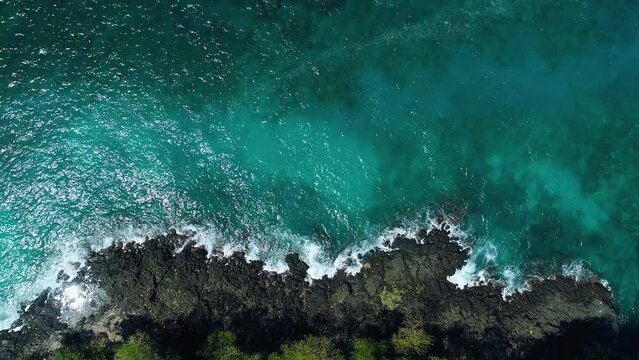 Top down aerial view on scenic light ocean waves breaking at the rocky shore with black volcanic lava rocks and dense jungle forest at the edge of the rainforest jungle landscape.