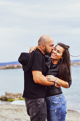 Portrait of happy    smiling  Couple standing on cliff against sea during  the winter .Young happy Bearded muscular  man  kissing and hugging beautiful woman  in leather jacket and hat on a beach