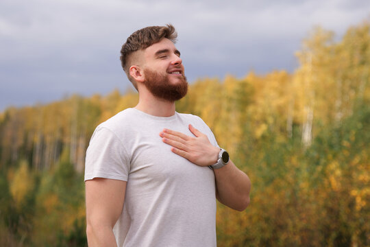 young bearded relaxed calm man enjoying good weather in the forest, autumn park, breathing deep fresh air and smiling. Guy with beard holding his hand on chest, heart with his eyes closed