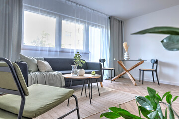 Spacious living room with window and curtains, comfortable furniture, sofa, armchair and table with chairs. Stylish room in modern interior with wooden floor and green plant.
