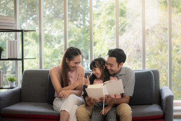 Obraz premium Asian father, mother and daughter Read a book and relax on the sofa in the living room and Happy on holidays and close together.