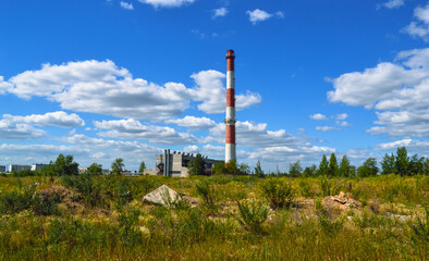 Fototapeta na wymiar A wild field and a tall striped red and white chimney of a boiler house against the background of white clouds in a blue sky