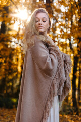 A very beautiful, confident woman in a brown poncho with closed eyes is enjoying autumn in the forest among yellow leaves.