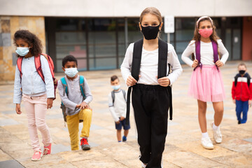 Confident teen girl in protective mask walking outside school building on spring day, going to lessons. Concept of necessary precautions in COVID pandemic.