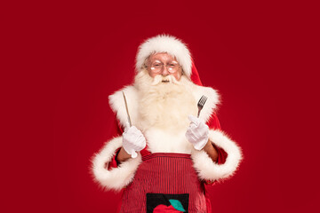 Christmas is coming ! Real Santa Claus holding fork and knife, looking at the camera with smile, posing over red studio background. Hungry, funny man.