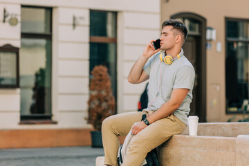 Fototapeta premium Young student guy sits on city street and speaks on mobile phone.