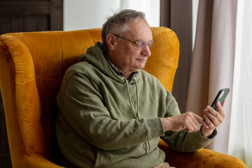 Senior male sits in armchair at home and using mobile phone