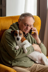 Senior male sits in armchair at home with a dog and talking by mobile phone