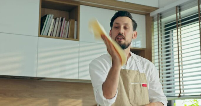 Selective focus on chef man in apron throwing in the air kneading homemade pizza dough in modern kitchen standing at wooden table relaxing while coking baking listening to music. Handsome husband .