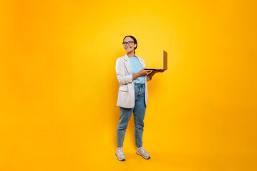 Full length photo of happy successful stylish latino or brazilian young woman, business employee, with glasses, hold in hands open laptop, stand on isolated orange background, looks to the side, smile