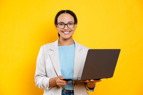 Positive confident clever lovely latino or brazilian young brunette woman, business lady, with glasses, hold in hands open laptop, stand on isolated orange background, looks at camera, smiles friendly