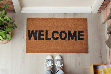 Woman standing near door mat with word Welcome on wooden floor in hall, top view - obrazy, fototapety, plakaty
