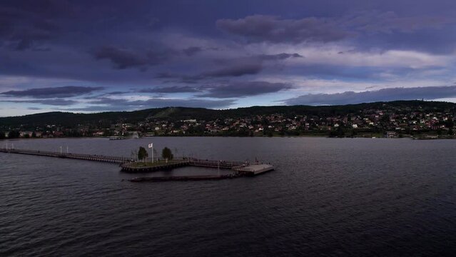 People walk on the wooden pier and boardwalk by a lake at sunset. Beautiful view of the town, mountains and blue purple sky with dramatic clouds during summer vacation. Sweden Aerial View 4K