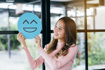 Asian woman holding blue happy smile face on paper cut, user giving good feedback rating, think...