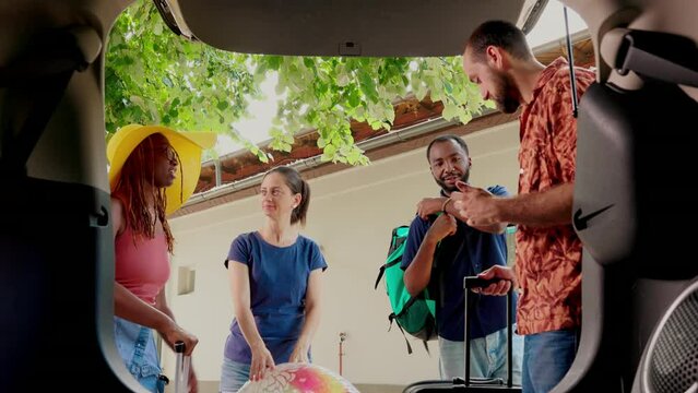Multiethnic friends putting voyage trolleys inside car trunk while getting ready for summer holiday citybreak. Diverse people going on vacation while loading vehicle with luggage.
