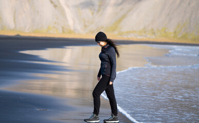 adventurous woman with cap and sunglasses walking on the black volcanic sand beach at the seashore...