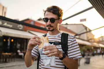 One man young adult modern caucasian male in the city in sunny day walk and eat sandwich fast food concept urban life copy space tourist eating on the street real person - Powered by Adobe