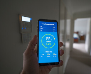 A man checks the charging status of a home installed battery storage unit on a smartphone and contemplates power consumption near an air conditioning control panel.