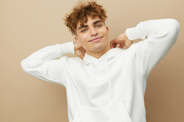 a happy, attractive man stands on a beige background in a light hoodie and smiling pleasantly looks at the camera with his hands behind his head