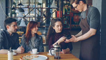 Young pretty woman is paying with smartphone for meal in cafe after lunch with friends then talking with mates. Modern technology, contactless payment and eating out concept.
