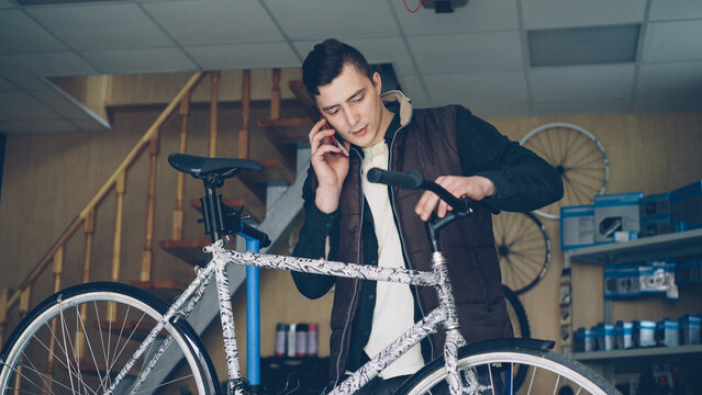 Attractive young man cheerful mechanic is busy talking on mobile phone and assembling bicycle handle-bar. People, maintenance and communication concept.