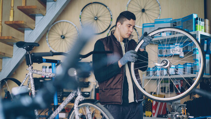 Experienced mechanic is rotating bicycle wheel checking work of internal mechanism and listening to music while working in small workshop. Maintenance and people concept.