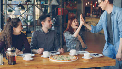 Handsome young guy in casual denim clothes is meeting his cheerful friends in cafe coming late and greeting his mates with high five. Friendship and eating out concept.