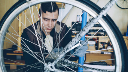 Concentrated handsome guy skilled mechanic is repairing bicycle wheel with wrenchwhile working in his workplace. Bike maintenance, people and profession concept.