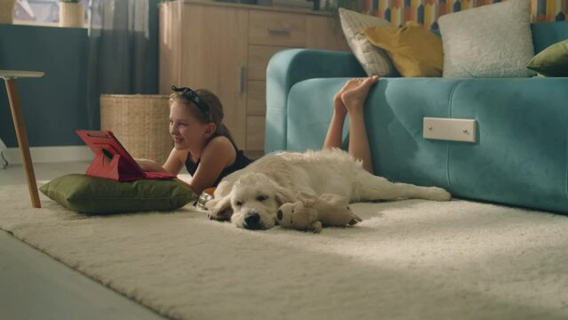 Young girl lying on mild carpet, choosing video on digital tablet computer, dog feeling tired and trying to sleep, spending leisure time at home with pet. Golden retriever.