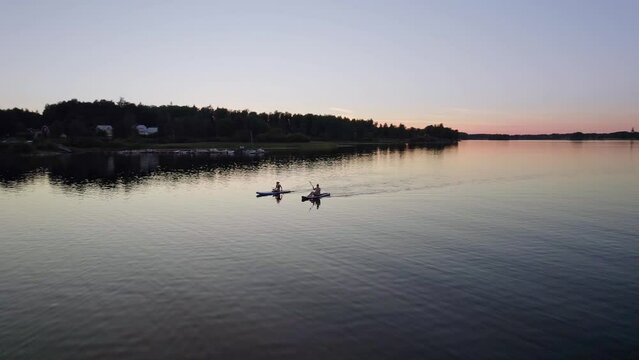 Young women paddle on a sup board on a lake towards the beach. Behind the two people a warm pink, orange sunset blue hour in the summer. Arc shot of an active summer vacation, Sweden on Lake Bysjön